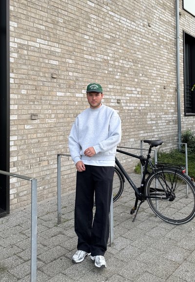 Sudadera gris, pantalones negros y zapatillas grises. Gorra verde con texto. Posicionada al lado de una bicicleta negra sobre una superficie pavimentada.
