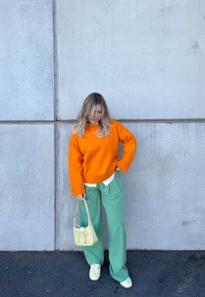 Mujer de pie contra una pared de hormigón, vistiendo un suéter naranja, pantalones verdes de pierna ancha, zapatos blancos y sosteniendo un bolso amarillo claro.