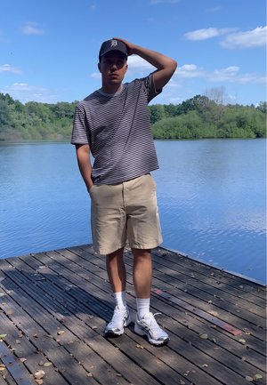 Camisa de manga corta a rayas, pantalones cortos beige, calcetines blancos y zapatillas; de pie en un muelle de madera junto a un lago con vegetación al fondo.