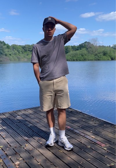 Camisa de manga corta a rayas, pantalones cortos beige, calcetines blancos y zapatillas; de pie en un muelle de madera junto a un lago con vegetación al fondo.