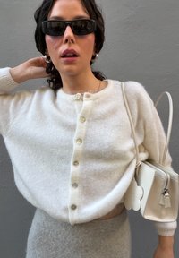 Valge soonikkoega kardigan ümmarguse kaelusega, viie nupuga, kantud halli rõiva peale; pehme tekstuur; aksessuaaridena mustad päikeseprillid ja valge käekott.