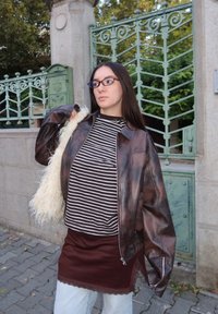 Hnědá kožená bunda, černobíle pruhované tričko, vínová sukně a světle modré džíny. Doplňky zahrnují chlupatou bílou tašku.