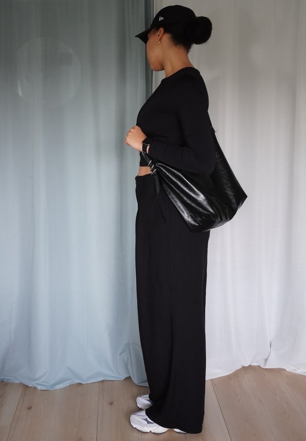 Noir top à manches longues, pantalon noir taille haute et sac noir brillant. Des baskets blanches complètent la tenue. L'arrière-plan présente des drapés en tissu doux.