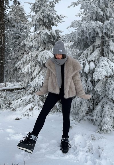Chaqueta de piel sintética en beige claro, leggings negros, gorro de punto gris, bufanda a juego, botas de invierno con acentos en negro y gris, fondo nevado.
