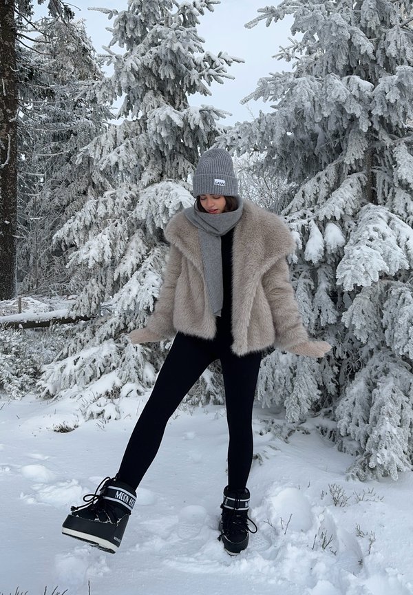 Kurtka z futra syntetycznego w jasnym beżu, czarne legginsy, szary dzianinowy beanie, pasujący szalik, zimowe buty z czarnymi i szarymi akcentami, śnieżne tło.