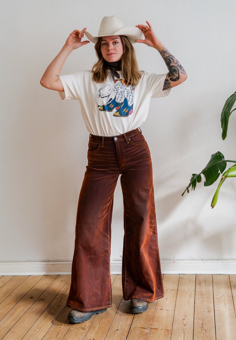 Ung kvinde med langt hår iført hvid cowboyhat, grafisk t-shirt, brune bukser med vide ben og støvler, står på trægulv ved siden af en grøn plante.