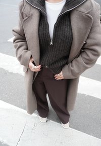 Manteau en laine marron sur un pull à fermeture éclair côtelé foncé, chemise blanche et pantalon large marron ; chaussures claires ; mains ornées de bagues.