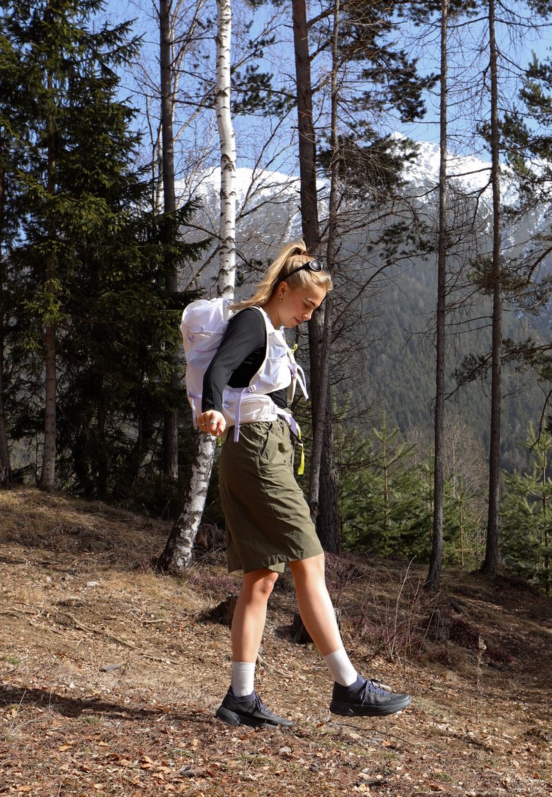Junge Frau wandert bergab auf einem Waldweg, trägt einen Rucksack, schwarzes Langarmshirt, grüne Shorts und schwarze Turnschuhe mit weißen Socken.