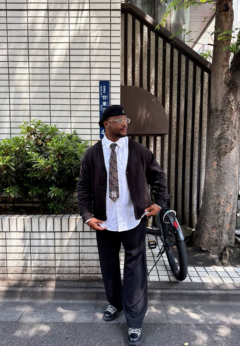 Homme portant un béret, des lunettes, une chemise blanche, une cravate à motifs et un cardigan noir, debout près d'un arbre et d'un vélo sur un trottoir en ville.