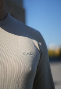 Beige dlouhý rukáv košile s ribovanou texturou. Má jemné logo na hrudi. Hladká tkanina, přiléhavý střih, na pozadí modré oblohy.