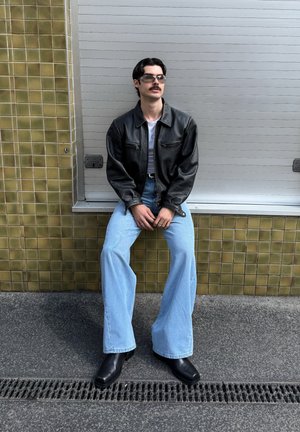 Jachetă din piele neagră, maiou alb, blugi largi de culoare albastru deschis și cizme negre până la gleznă. Accesoriile includ ochelari de soare. Fundalul prezintă plăci cu textură.