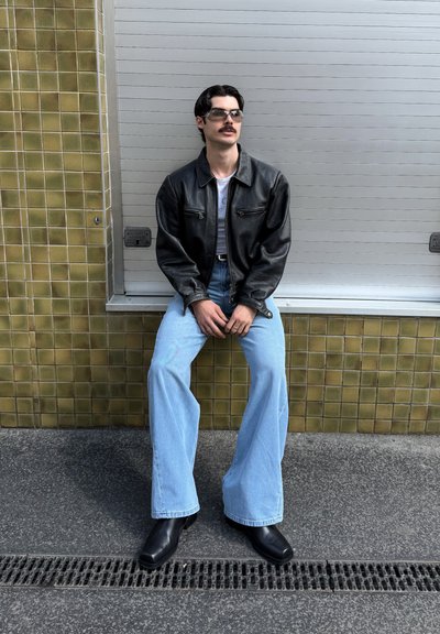 Chaqueta de cuero negra, camiseta blanca, jeans de pierna ancha azul claro y botas negras de tobillo. Los accesorios incluyen gafas de sol. El fondo presenta baldosas texturizadas.