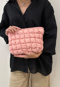 Pochette rose rembourrée avec un design matelassé, dotée d'un bord froncé. Tenue par une personne portant une chemise noire oversized et un pantalon clair.