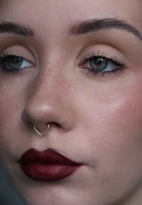 Close-up van een gezicht met een gouden hartvormige neusbella, donkerrode matte lippen, lichte huid en groene ogen met subtiele make-up.