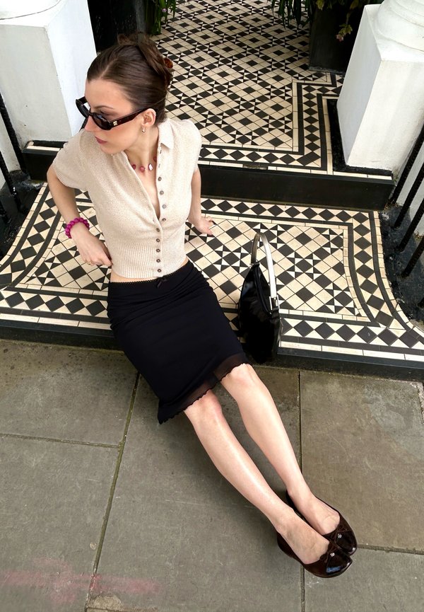 Femme portant des lunettes de soleil, un haut beige et une jupe noire, assise sur une marche carrelée noir et blanc à côté d'un sac à main noir sur un sol en pierre.
