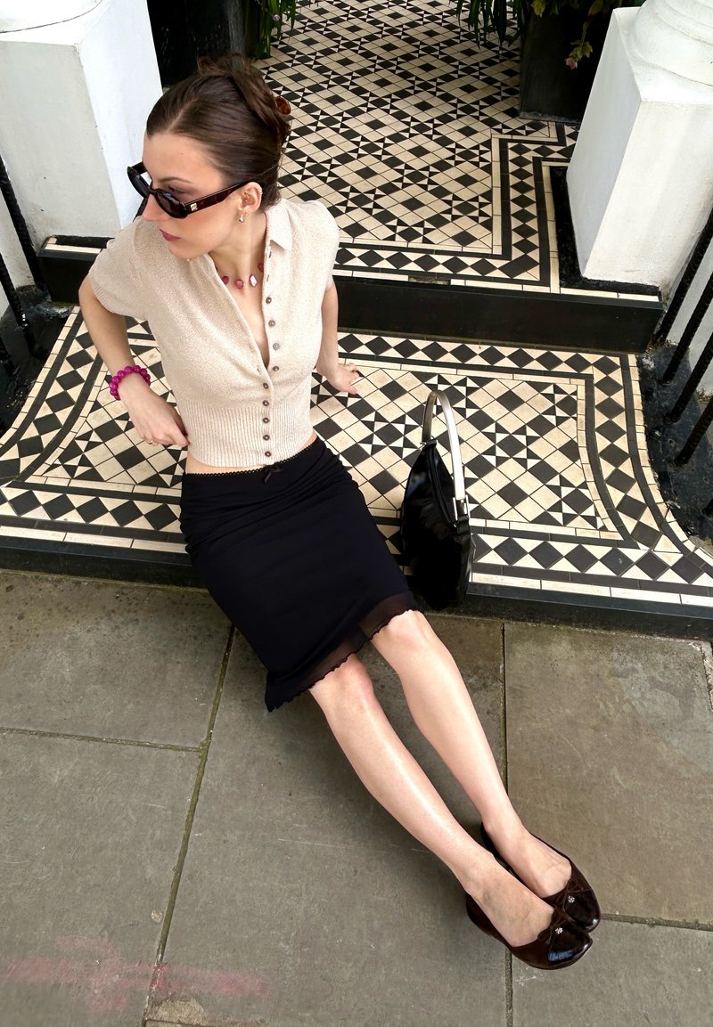 Femme en lunettes de soleil portant un haut beige et une jupe noire assise sur une marche carrelée noir et blanc à côté d'un sac à main noir sur un pavé en pierre.