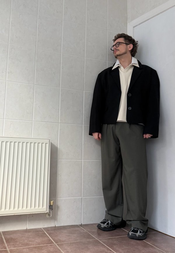 Un homme portant des lunettes, une veste noire, une chemise blanche et un pantalon ample se tient près d’un mur carrelé et d’un radiateur.