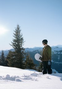 Snowboarder en sweat-shirt marron et pantalon noir, tenant un snowboard blanc avec des lettres noires, devant un paysage de montagnes enneigées.