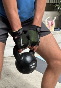 Melnā kettlebell ar uzrakstu "ALEX 12KG", turēta abām rokām, uzvilktas melnas un zaļas bezpirkstu cimdi, kokvilnas šorti un zila T-krekls.
