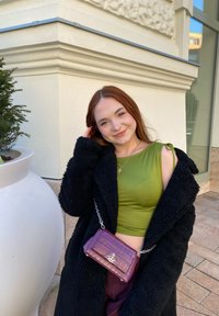 Πράσινη αμάνικη μπλούζα, μαύρο φουντωτό παλτό και μια μικρή τσάντα χεριού με μοτίβο κροκοδείλου σε μωβ χρώμα με ασημί λεπτομέρειες. Ουδέτερο φόντο με φυτά.