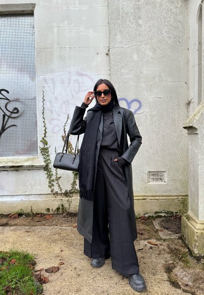 Mujer con abrigo de cuero negro, pantalones de pierna ancha, bufanda, gafas de sol y bolso, de pie frente a una pared con grafiti y hiedra.