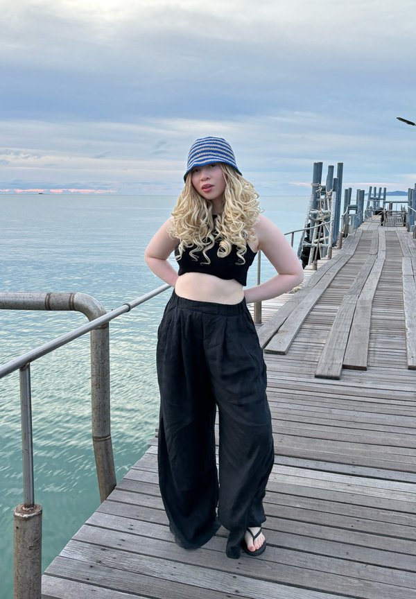 Calças largas pretas e uma blusa preta cropped, complementadas por um chapéu de balde listrado em azul e branco, estão em pé num cais de madeira junto à água.