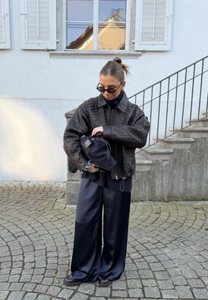 Mujer con chaqueta de cuero negra tejida y pantalones anchos negros ajustando un bolso negro al aire libre junto a escalones de piedra y una ventana.