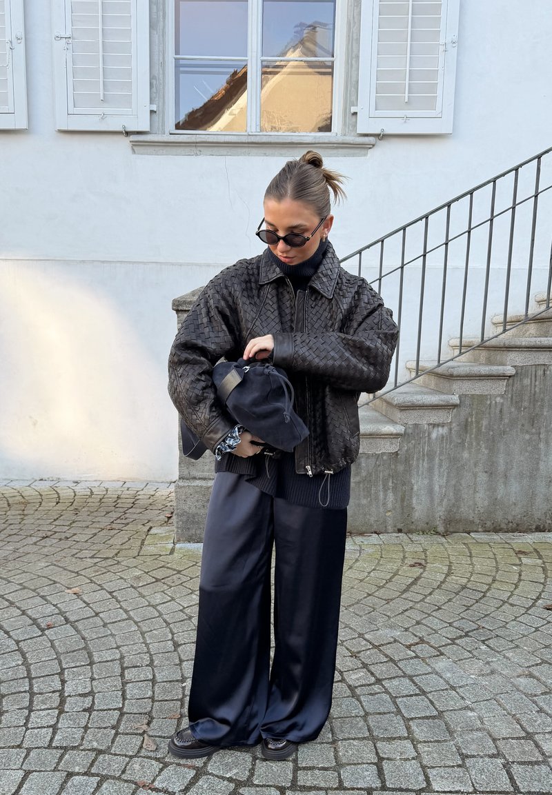 Femme en veste en cuir tressé noire et pantalon large noir ajustant un sac noir à l'extérieur près de marches en pierre et d'une fenêtre.