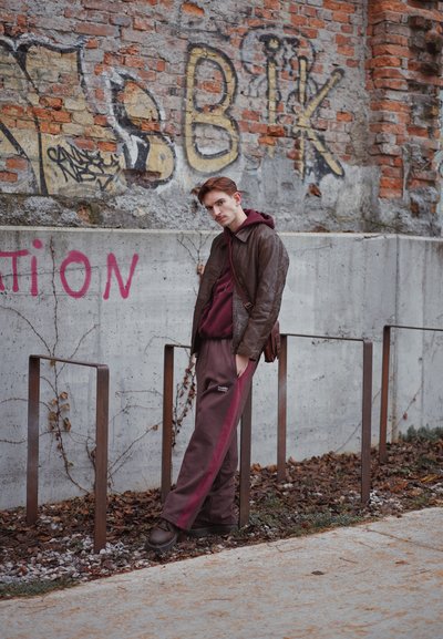 Chaqueta de cuero marrón, sudadera con capucha color granate y pantalones marrones con franjas granate. Está junto a postes de metal contra una pared de ladrillos texturizada.