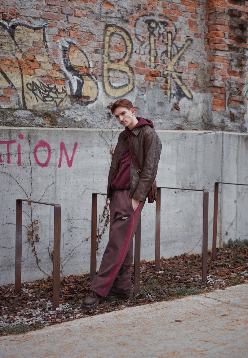 Braune Lederjacke, dunkelroter Kapuzenpullover und braune Hose mit dunkelroten Streifen. Steht neben Metallpfosten vor einer strukturierten Backsteinmauer.