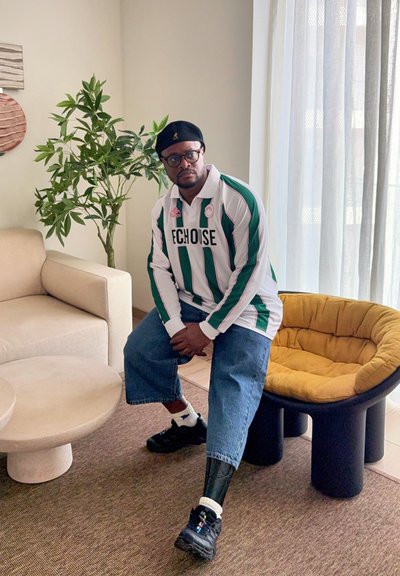 Hombre con boina negra, gafas, camiseta de rayas verdes y blancas y pantalones vaqueros recortados sentado en una silla amarilla acolchada en una sala de estar.