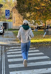 Pessoa vestindo uma jaqueta bege, jeans azuis e um boné, atravessando uma faixa de pedestres ao lado de carros estacionados e árvores de outono.