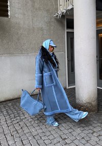 Μακρύ μπλε παλτό με ανοιχτό μπλε περιθώριο, συνδυασμένο με ρέουσες τζιν παντελόνες και μπλε αξεσουάρ. Μία δομημένη τσάντα συμπληρώνει το ντύσιμο.