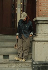 Jeansjakke, grå strikket lue, oversized beige bukser og beige slip-on sko. Liten mønstret håndveske og lagdelte halskjeder. Murvegg i bakgrunnen.