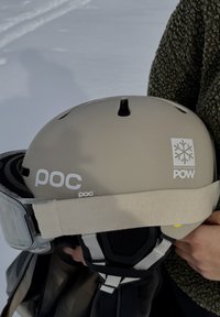 Φωτεινό καφέ κράνος σκι POC με γυαλιά σκι κρατημένα απέναντι από ένα άτομο που φοράει πράσινο φλις μπουφάν σε χιονισμένο περιβάλλον.