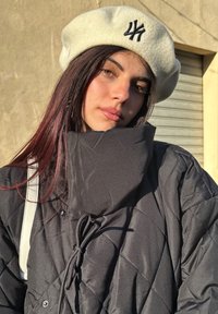 Czarna pikowana kurtka z wysokim kołnierzem, w połączeniu z kremowym beretem ozdobionym czarnym haftowanym logo. Włosy falowane z bordowymi refleksami.