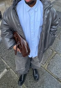 Doudoune grise, chemise boutonnée bleu clair, jeans gris, bottines noires. Tient un sac en cuir marron avec des détails métalliques.