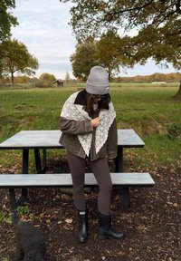 Brown leather jacket with a black collar, layered with a white lace scarf. Dark leggings and black rubber boots. Outdoor setting with a picnic table.