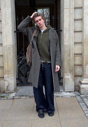 Una persona lleva un abrigo largo de cuadros gris sobre una sudadera con capucha verde, combinado con pantalones anchos de mezclilla oscura y zapatos negros. Lleva una bolsa marrón colgada de un hombro.