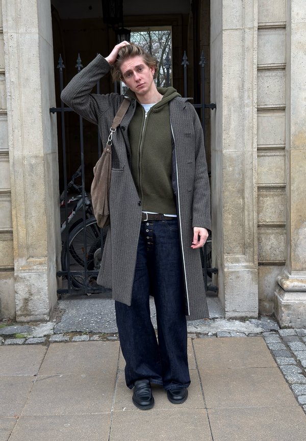 Inimene kannab pikkade varrukatega halli ruudulist mantlit rohelise kapuutsiga pusa kohal, seljas on laiad tumedad teksapüksid ja jalas mustad jalatsid. Ühe õla peal ripub pruun kott.