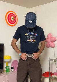 Mand iført en marineblå kasket, en marineblå grafisk t-shirt med nautiske flag, brune bukser og flere ringe og halskæder i et farverigt rum.
