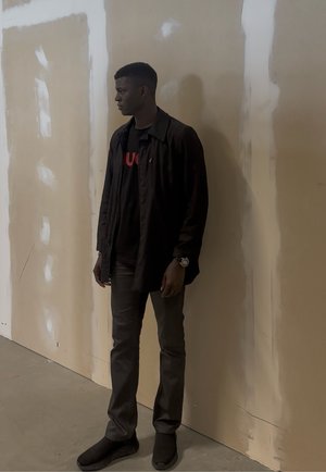 Joven con chaqueta negra, pantalones oscuros y zapatos, de pie mirando hacia la izquierda contra una pared beige con manchas blancas.