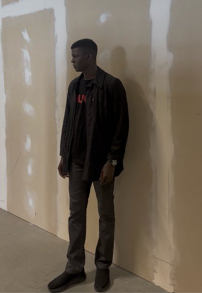 Joven con chaqueta negra, pantalones oscuros y zapatos, de pie mirando hacia la izquierda contra una pared beige con manchas blancas.