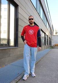 Sarkans un jūras spēku zilas krāsas sporta krekls ar apļu logotipu „90”, uzvilkts virs melna garām piedurknēm krekla, saskaņots ar gaiši ziliem džinsiem un pelēkiem sporta apaviem.
