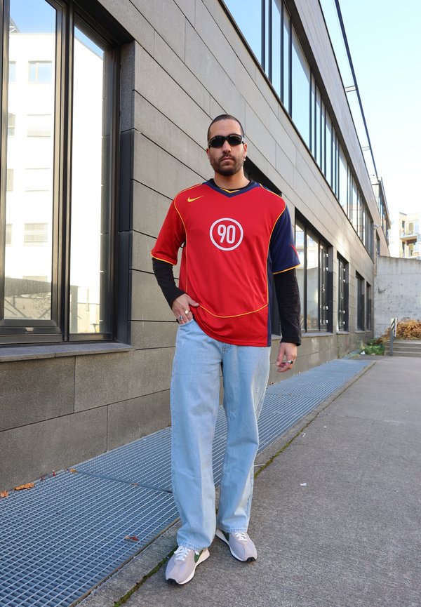 Red and navy athletic jersey with a circular ‘90’ logo, worn over a black long-sleeve shirt, paired with light blue jeans and gray sneakers.