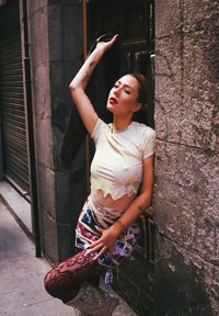 Durchsichtiger, hellgelber Oberteil mit wellenförmigem Saum; gemusterter, hochtaillierter Rock in lebendigen Farben; dunkle Spitzenstrumpfhose; silberne Plateau-Stiefel.