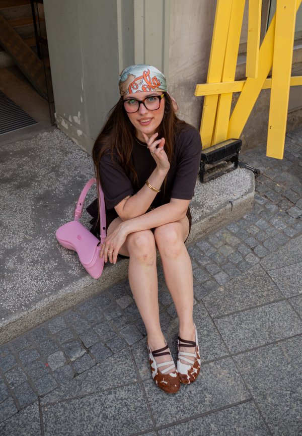 Jonge vrouw met bril en een patroon hoofddoek zit op een stoeprand, draagt een zwarte jurk en tweekleurige ballerina's, naast een roze handtas.