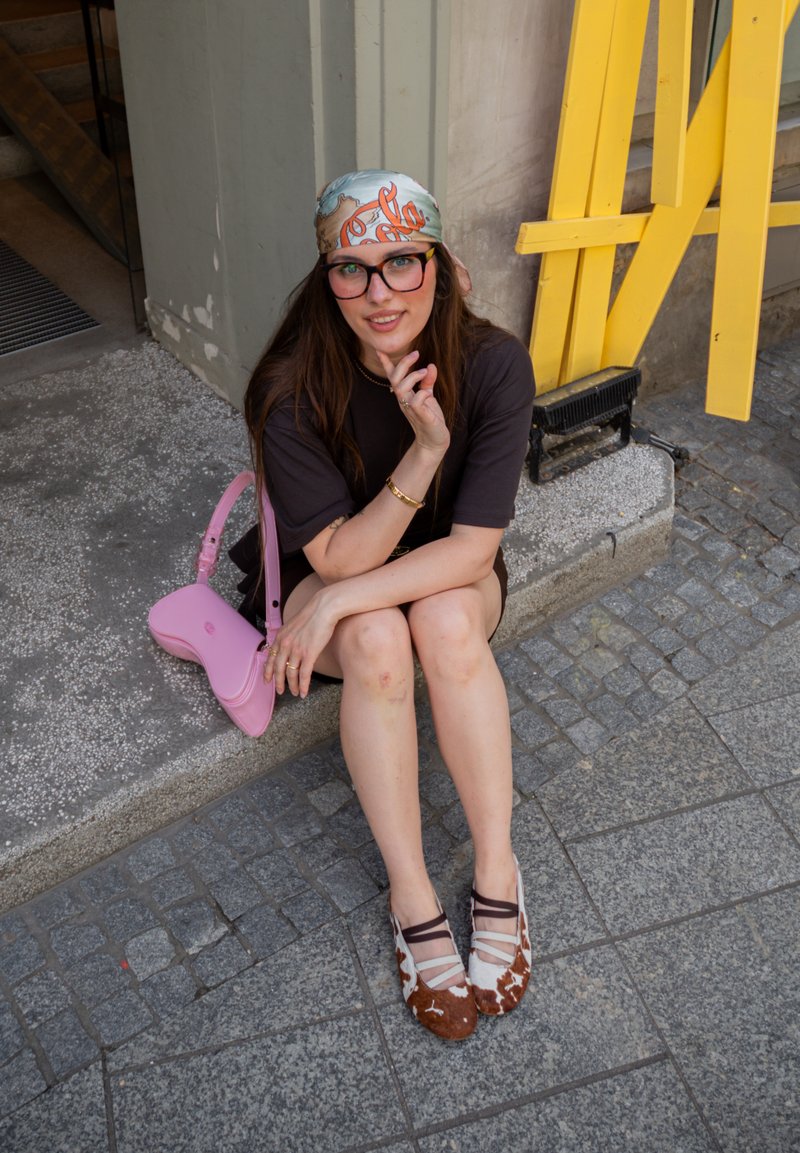 Jonge vrouw met bril en een patroon hoofddoek zit op een stoeprand, draagt een zwarte jurk en tweekleurige ballerina's, naast een roze handtas.