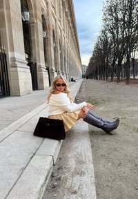 Brun semsket skinnveske, kremfarget genser, gyllen skjørt og grå knelange støvler. Bakgrunnen viser en kolonnade og trær som fyller en sti.