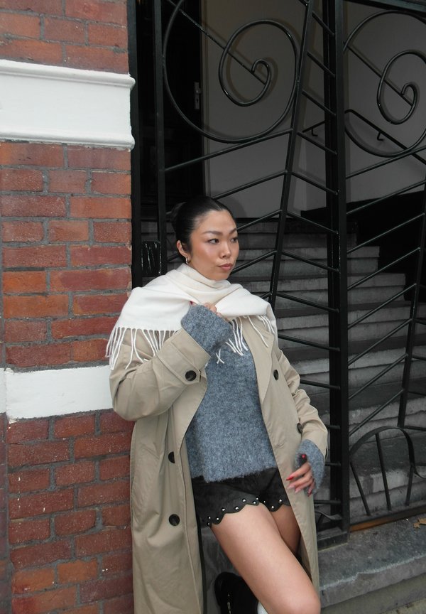 Beige trenchcoat, grå genser, svarte shorts med bølgete kanter, hvitt skjerf med frynser. Bakgrunnen har teksturert murstein og smijernsrekkverk.